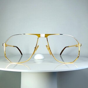 May include: Gold-rimmed aviator eyeglasses with a double bridge and clear lenses. The frames have a gold finish with silver accents on the top bar and temples. The glasses are displayed on a white, round pedestal.