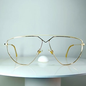 Könnte beinhalten: Übergroße Brille im Pilotenstil mit einem dünnen, polierten goldfarbenen Metallrahmen. Das Design umfasst einen markanten Doppelsteg an der Oberseite, klare Gläser und schlanke Bügel mit gelblichen Enden für eine leichte Optik.