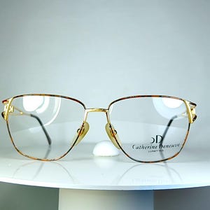 Può includere: Occhiali dorati con motivo tartarugato marrone sui bordi superiori e sulle aste. Le lenti sono trasparenti e la montatura presenta la scritta "Catherine Deneuve Lunettes". Gli occhiali poggiano su una superficie bianca.