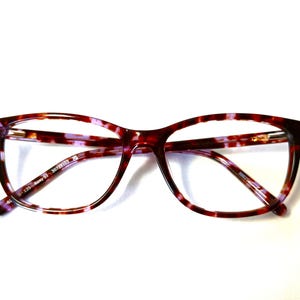 Peut inclure: Une paire de lunettes avec un motif &eacute;caille de tortue dans les tons de rouge, marron et violet. Les montures sont de forme rectangulaire avec des bords arrondis. Les verres sont clairs et les branches sont fines.