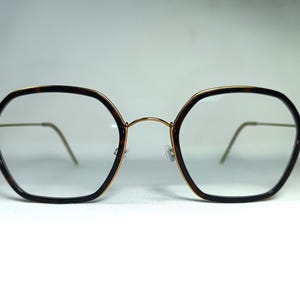 Może przedstawiać: Okulary z oprawkami w wzór szylkretu i złotymi metalowymi zausznikami. Soczewki są przezroczyste, a oprawki mają unikalny ośmiokątny kształt. Okulary są na białym tle.