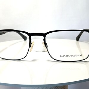 May include: Black rectangular eyeglasses with clear lenses. The frames are made of metal and feature the "Emporio Armani" logo on the right lens. The glasses are set on a white surface.