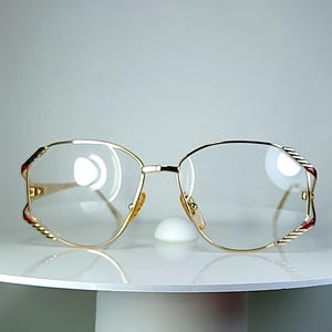 Könnte beinhalten: Goldrandbrille mit einzigartigem Design. Die Fassungen haben eine goldene Oberfläche mit roten und weißen Akzenten an den Bügeln. Die Gläser sind klar, und die Brille wird auf einer weißen Oberfläche präsentiert.