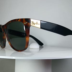 May include: Tortoiseshell sunglasses with dark green lenses and black temples. The sunglasses have a gold accent with the brand name. The sunglasses are resting on a white surface.