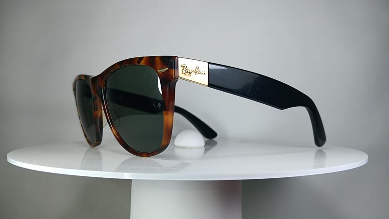 May include: Tortoiseshell sunglasses with dark green lenses and black temples. The sunglasses have a gold accent with the brand name. The sunglasses are resting on a white surface.