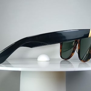 May include: Black sunglasses with a tortoiseshell pattern frame and green lenses. The sunglasses have a gold accent on the temples and are resting on a white surface. A small, white, dome-shaped object is also on the surface.