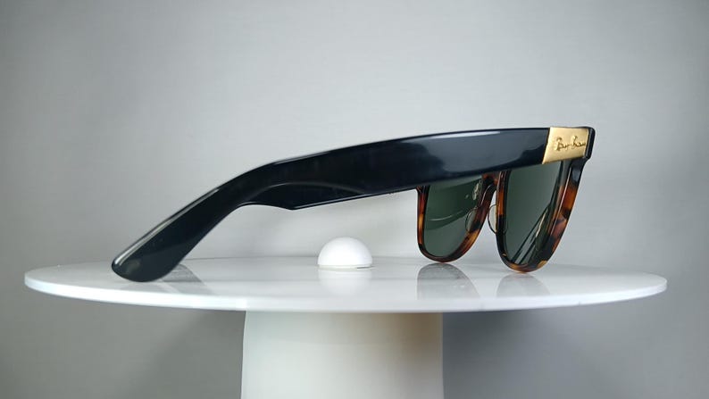 May include: Black sunglasses with a tortoiseshell pattern frame and green lenses. The sunglasses have a gold accent on the temples and are resting on a white surface. A small, white, dome-shaped object is also on the surface.