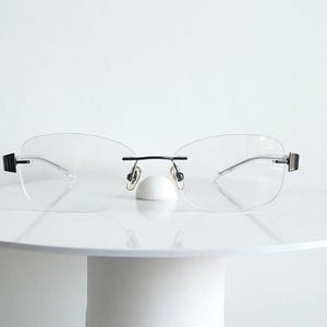 以下が含まれることがあります： 透明なレンズ、細い黒のブリッジ、黒のテンプルを備えたリムレスメガネ。調整可能なノーズパッドとミニマルな構造を持ち、ヒンジ部分にシルバー調のアクセントが施された洗練されたデザインが特徴です。