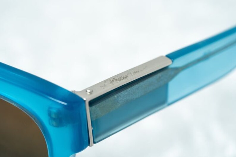 K&ouml;nnte beinhalten: Nahaufnahme einer blauen Sonnenbrille mit einem silbernen Metallgelenk. Das Gelenk tr&auml;gt die Gravur "Made in Italy".