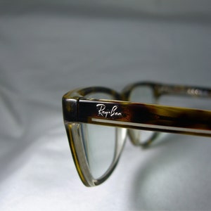 May include: Close-up of a pair of tortoiseshell eyeglasses with a silver Ray-Ban logo on the frame.