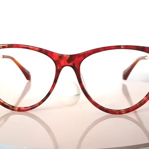 May include: Red and brown tortoiseshell eyeglasses with a cat-eye shape. The frames have a speckled pattern and gold-colored arms. The lenses are clear, and the glasses are displayed on a white surface.