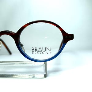 May include: Round eyeglasses with a brown and blue gradient frame. The glasses have a classic design with the text "BRAUN CLASSICS" printed on the lens. The frame is supported by a clear stand.