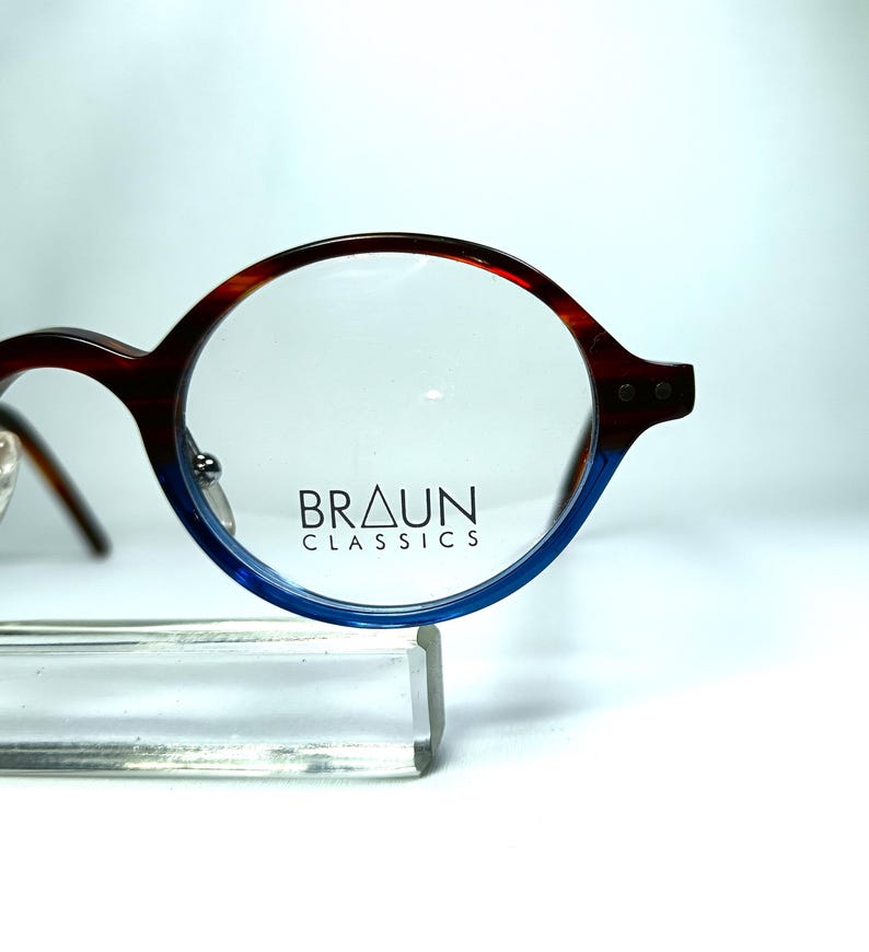 May include: Round eyeglasses with a brown and blue gradient frame. The glasses have a classic design with the text "BRAUN CLASSICS" printed on the lens. The frame is supported by a clear stand.