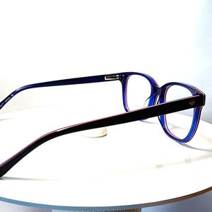 May include: A pair of eyeglasses with a dark blue frame and temples, and a black bridge. The lenses are clear. The glasses are resting on a white surface.