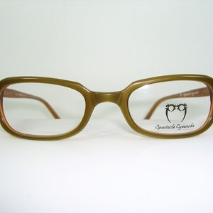 Puede incluir: Gafas rectangulares de color verde oliva con lentes transparentes. El nombre de la marca "Spectacle Eyeworks" está impreso en la lente derecha.