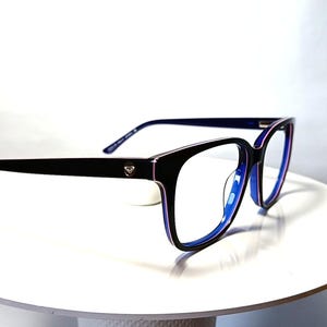May include: Black-framed eyeglasses with a square shape and blue accents. The glasses have a small decorative emblem on the temples. The lenses are clear, and the frame is set against a white background.