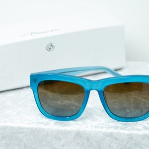 K&ouml;nnte beinhalten: Blaue quadratische Sonnenbrille mit braunen Gl&auml;sern. Die Sonnenbrille liegt auf einer wei&szlig;en, flauschigen Oberfl&auml;che.