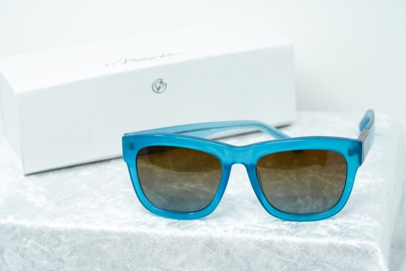 K&ouml;nnte beinhalten: Blaue quadratische Sonnenbrille mit braunen Gl&auml;sern. Die Sonnenbrille liegt auf einer wei&szlig;en, flauschigen Oberfl&auml;che.