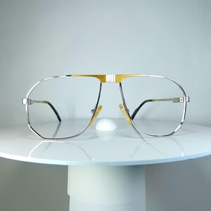 May include: Silver and gold-toned aviator eyeglasses with clear lenses. The glasses have a double bridge and a gold accent on the top bar. The temples are thin and black. The glasses are displayed on a white surface.