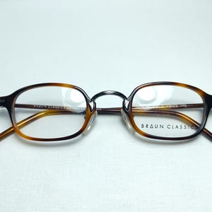 May include: Brown and tortoise shell eyeglasses with clear lenses. The glasses have a classic oval shape and a metal bridge. The brand name "BRAUN CLASSIC" is printed on the right lens. The glasses are set against a white background.