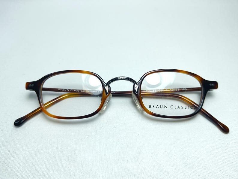 May include: Brown and tortoise shell eyeglasses with clear lenses. The glasses have a classic oval shape and a metal bridge. The brand name "BRAUN CLASSIC" is printed on the right lens. The glasses are set against a white background.