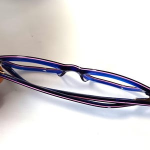 May include: A pair of eyeglasses with a black frame, accented with a thin pink stripe along the top and blue on the inside. The lenses are clear, and the glasses are set against a white background.