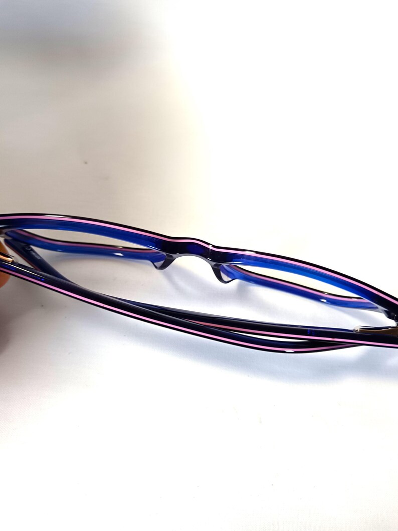 May include: A pair of eyeglasses with a black frame, accented with a thin pink stripe along the top and blue on the inside. The lenses are clear, and the glasses are set against a white background.