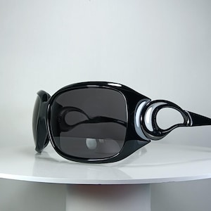 May include: Oversized black sunglasses featuring a glossy finish and dark tinted lenses. The frames have a distinct, rounded shape with a unique cutout design on the temples, creating a bold look with smooth, curved lines throughout the entire structure.