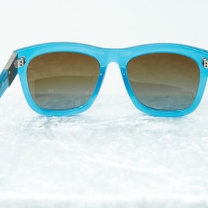 K&ouml;nnte beinhalten: Blaue Kunststoff-Sonnenbrille mit braunen Verlaufsgl&auml;sern. Die Sonnenbrille hat einen d&uuml;nnen Metallrahmen und befindet sich auf einem wei&szlig;en Hintergrund.