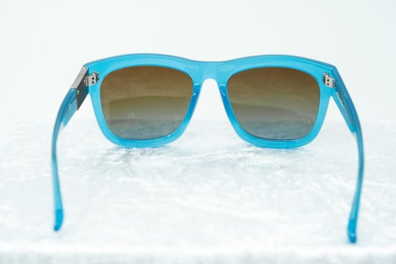 K&ouml;nnte beinhalten: Blaue Kunststoff-Sonnenbrille mit braunen Verlaufsgl&auml;sern. Die Sonnenbrille hat einen d&uuml;nnen Metallrahmen und befindet sich auf einem wei&szlig;en Hintergrund.