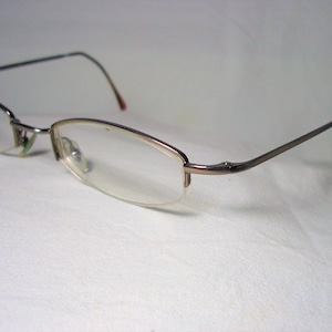 May include: A pair of silver-toned eyeglasses with clear lenses. The glasses have a thin, rectangular frame and are resting on a white surface.