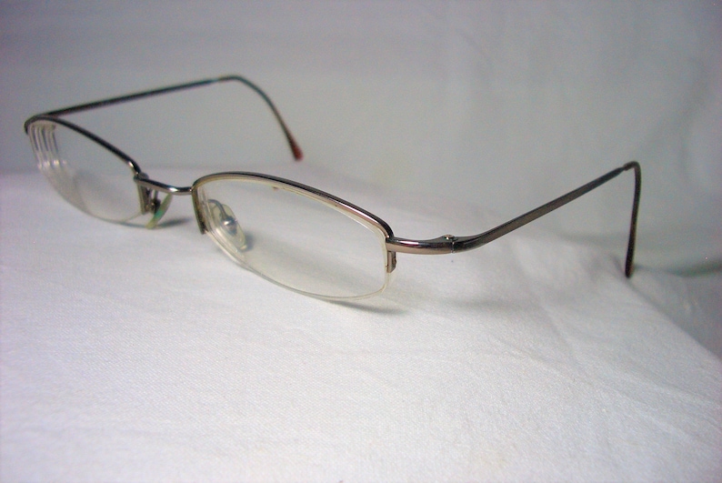 May include: A pair of silver-toned eyeglasses with clear lenses. The glasses have a thin, rectangular frame and are resting on a white surface.
