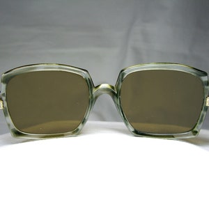 Może przedstawiać: Okulary przeciwsłoneczne z zielonego plastiku z brązowymi szkłami. Okulary mają kwadratowy kształt i są ostre.