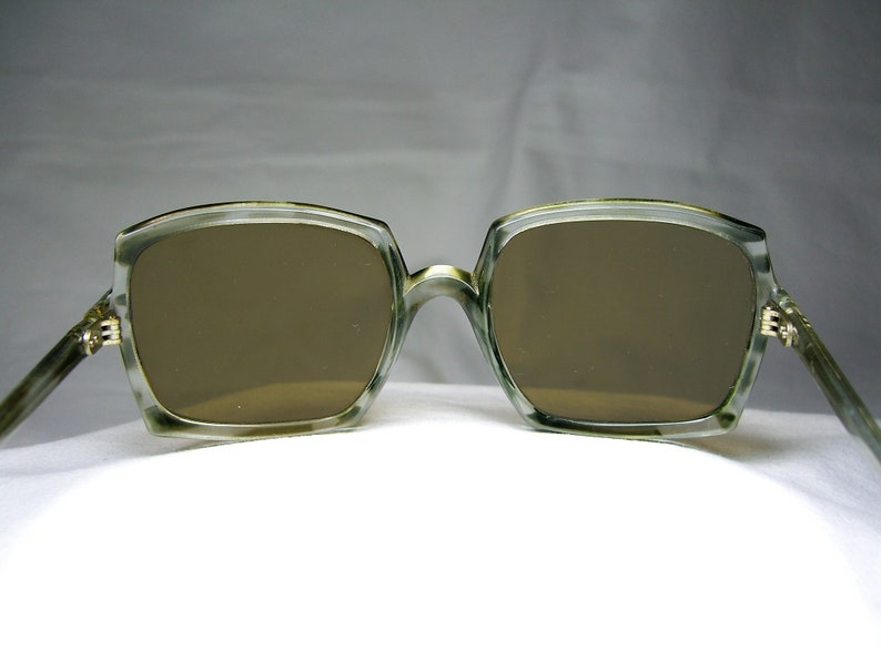 Może przedstawiać: Okulary przeciwsłoneczne z zielonego plastiku z brązowymi szkłami. Okulary mają kwadratowy kształt i są ostre.