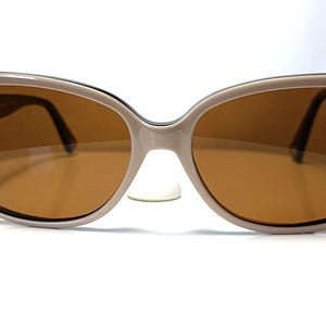 May include: Oversized sunglasses with a light brown frame and brown lenses. The sunglasses have a rounded, slightly cat-eye shape. The lenses are a warm brown color, and the frame is a neutral beige. The sunglasses are set against a white background.