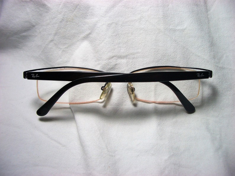 May include: Black rectangular eyeglasses with a thin metal frame and clear lenses. The brand name "Ray-Ban" is visible on the right temple.
