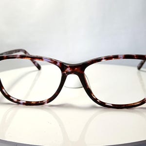 Peut inclure: Lunettes &agrave; motif &eacute;caille de tortue. Les montures sont un m&eacute;lange de marron, de noir et de blanc, de forme rectangulaire. Les verres sont clairs. Les lunettes sont pos&eacute;es sur une surface blanche.
