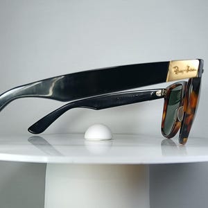 May include: Black sunglasses with a tortoiseshell pattern and green lenses. The sunglasses have a gold accent on the temple and are resting on a white surface. The Ray-Ban logo is visible on the temple.