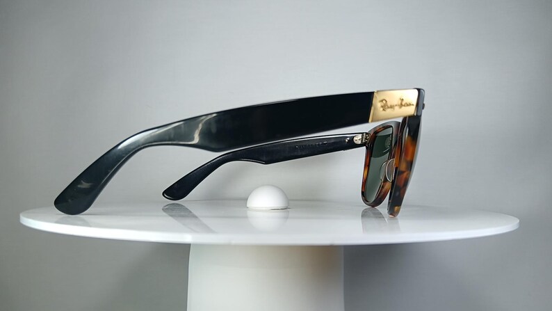 May include: Black sunglasses with a tortoiseshell pattern and green lenses. The sunglasses have a gold accent on the temple and are resting on a white surface. The Ray-Ban logo is visible on the temple.