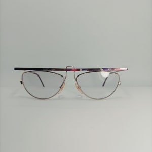Peut inclure: Une paire de lunettes à monture dorée avec un pont unique à motifs multicolores. Les verres sont transparents et les lunettes ont une forme légèrement œil de chat.