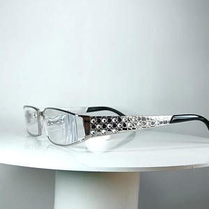 May include: Silver-toned spectacles featuring clear lenses and a rectangular frame. The temples are embellished with a grid of small, sparkling clear rhinestones, transitioning into smooth black plastic tips at the ends for a secure fit.