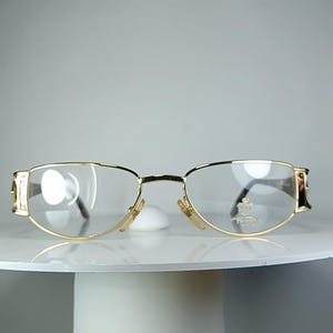 Könnte beinhalten: Goldrandbrille mit klaren Gläsern. Die Brille hat eine runde Form und wird auf einer weißen Oberfläche präsentiert. Die Bügel weisen dekorative Akzente auf. Das Gesamtdesign ist klassisch und elegant.