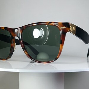 May include: Tortoiseshell sunglasses with dark green lenses. The frames have a classic design with a brown and black pattern. The temples are black with a gold accent. The sunglasses are displayed on a white surface.