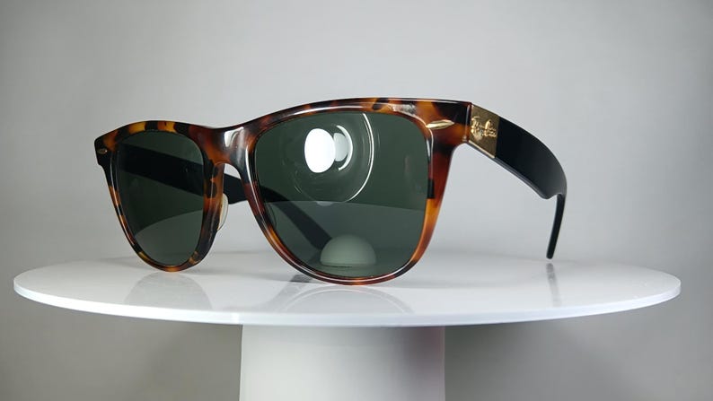 May include: Tortoiseshell sunglasses with dark green lenses. The frames have a classic design with a brown and black pattern. The temples are black with a gold accent. The sunglasses are displayed on a white surface.