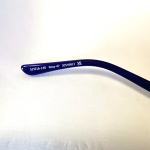 May include: Close-up of a dark blue Roxy eyeglass arm. The arm features the text "52▢16-140 Roxy 47 30549821 UK CA" printed in white. The arm curves slightly, with a rounded end.