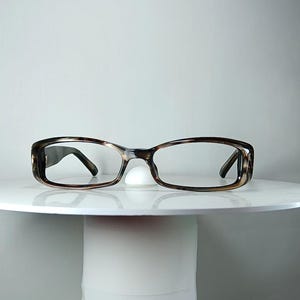 May include: Spectacles featuring a rectangular frame with a brown and black tortoiseshell pattern. The frame has a thin bridge and slim temples that extend outward from the sides, showcasing a classic design with a polished finish and clear lenses.