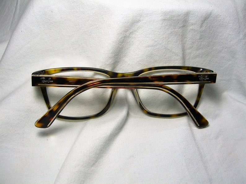 May include: Tortoise shell eyeglasses with a thin gold frame and clear lenses. The brand name "Ray-Ban" is visible on the right temple.