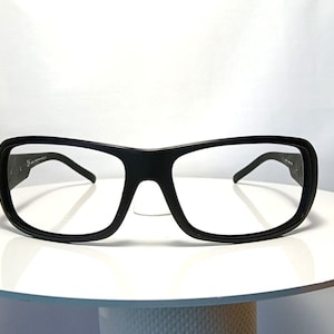 May include: Black rectangular-framed spectacles. The glasses have a thick, matte black frame and clear lenses. The temples are thin and dark. The spectacles are displayed on a white surface against a white background.