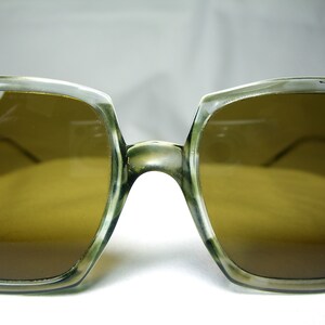 Może przedstawiać: Okulary przeciwsłoneczne vintage z jasnobrązową oprawką i brązowymi szkłami. Oprawka wykonana jest z przezroczystego plastiku i ma kwadratowy kształt.