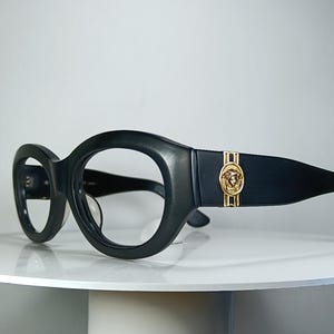 May include: Black eyeglasses with thick, rounded frames and wide temples. A circular gold-toned emblem featuring a Medusa head design is embedded on the side of each temple, accented by vertical gold-toned stripes on the top and bottom edges.